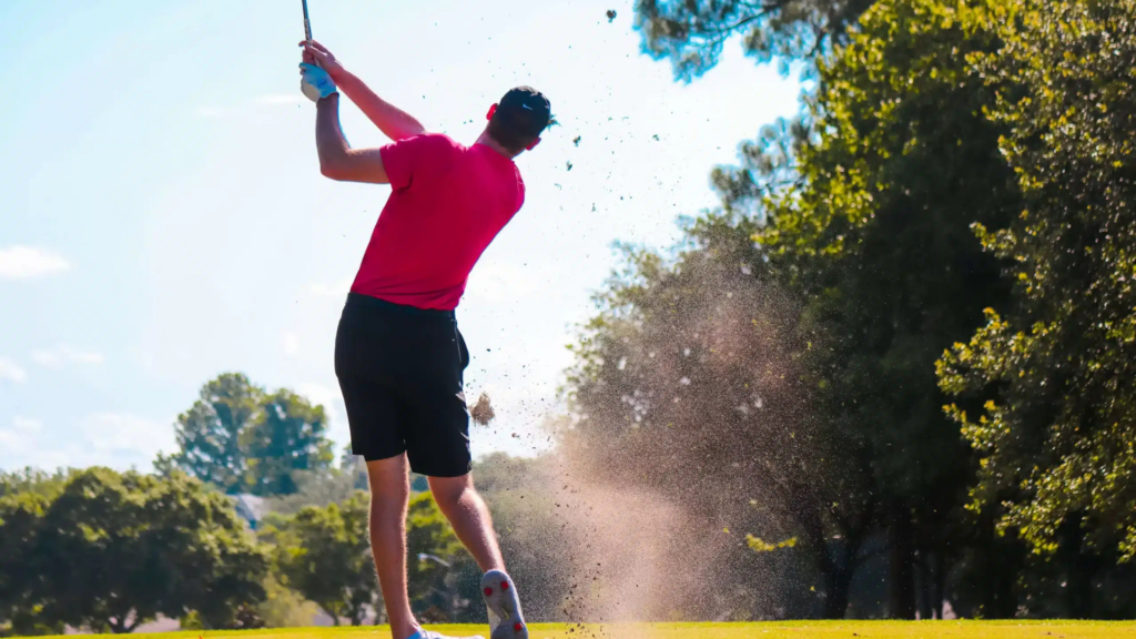 Golfer in pink shirt swinging club, dirt flying on sunny Chibana Golf course.